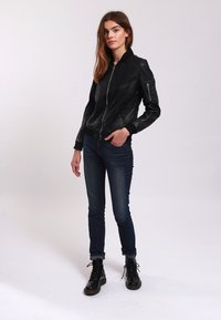 Schwarze Leder-Bomberjacke mit hohen Kragen, gerippten Manschetten und Reißverschluss vorne, getragen über dunklen Skinny-Jeans und schwarzen Stiefeln.
