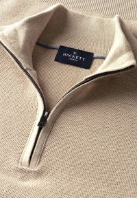 Béžový pletený sveter s štvrťzipsom a rebrovaným golierom, vo vnútri goliera je čierna štítka "Hackett London".