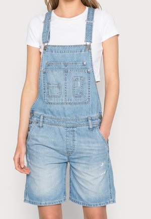 Salopette en jean bleu clair avec bretelles réglables, poches avant et détails usés ; portée avec un t-shirt court blanc.