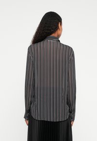 Vrouw met lang, krullend haar die een zwart, wit en grijs verticaal gestreept lange mouwen shirt en een zwarte plissérok aan heeft, met de rug naar de kijker gekeerd.