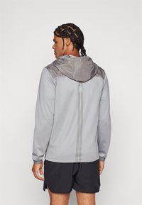 Svijetlo siva sportska jakna s tamnijim sivim kapuljačom. Ima jednostavan dizajn, duge rukave i vertikalni uzorak pruga na leđima.