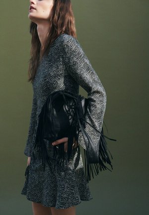 Ženska v obleki z dolgimi rokavi s potiskom, ki drži črno usnjeno torbico s cofki na enobarvni olivno zeleni podlagi.