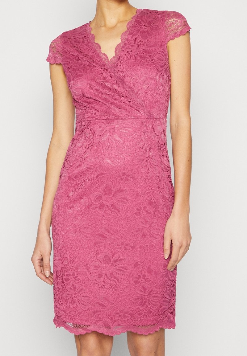 Robe ajustée en dentelle rose longueur genou avec manches courtes, encolure en V et motifs floraux portée par une personne debout.