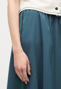 Detailní záběr osoby nosící krémový top a vysokým pasem tyrkysovou sukni se zipem po straně, paže volně podél těla.