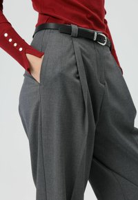 Grijze op maat gemaakte geplooide broek van een getextureerde stof, voorzien van een zwarte leren riem met een metalen gesp en zijzakken. Rode top met lange mouwen zichtbaar.