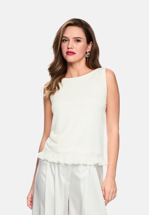 Woman with loose wavy hair wearing sleeveless white top with ruffled hem and white wide-leg pants, standing against plain background.