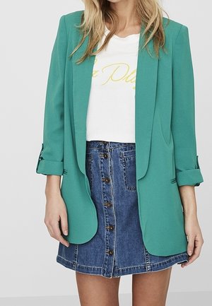 Kvinde iført teal blazer med opsmøgede ærmer, hvid grafisk t-shirt og denim nederdel med knapper foran, vist fra hals til midt på låret.