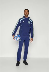 Mann i blå Adidas treningsdrakt som holder en blå og hvit fotball, stående mot en ensfarget lys grå bakgrunn.