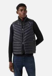 Czarna pikowana kamizelka z wysokim kołnierzem, zapięciem na zamek i bocznymi kieszeniami, ozdobiona wzorem w jodełkę i logo marki na piersi.