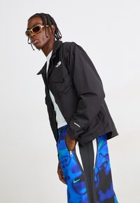 Mand med fletninger iført solbriller, en sort North Face jakke, hvid skjorte og blå mønstrede Nike bukser med hænderne i lommerne.