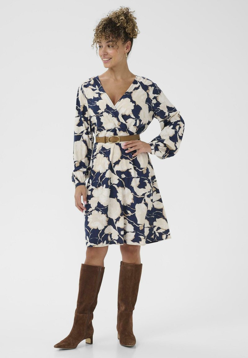 Wikkeljurk met bloemmotief in marineblauw en crèmekleurig, met volumineuze mouwen, getailleerd met een bruine riem. Bruine suède laarzen tot aan de knie.