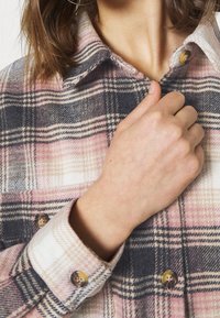 Camicia di flanella in colore rosa, grigio e beige con motivo a quadri. Presenta un colletto classico e chiusura a bottoni, con polsini arrotondati e una texture liscia.