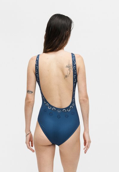 Woman with long dark hair wearing a blue one-piece swimsuit with decorative patterns and a low back, viewed from behind on a plain background.