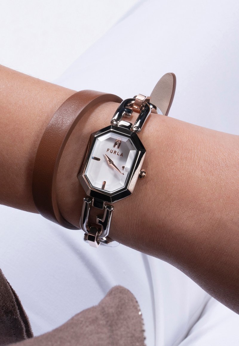 Furla watch with an octagonal silver-tone case, white dial, rose gold accents, linked bracelet, and a brown leather band on a wrist.