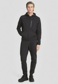 Felpa nera con cappuccio e pantaloni coordinati, realizzati in tessuto morbido. La giacca presenta una tasca con zip e i pantaloni hanno polsini elasticizzati. Accentuati da un cappuccio.