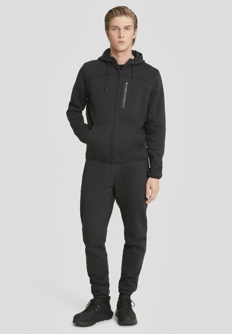 Felpa nera con cappuccio e pantaloni coordinati, realizzati in tessuto morbido. La giacca presenta una tasca con zip e i pantaloni hanno polsini elasticizzati. Accentuati da un cappuccio.