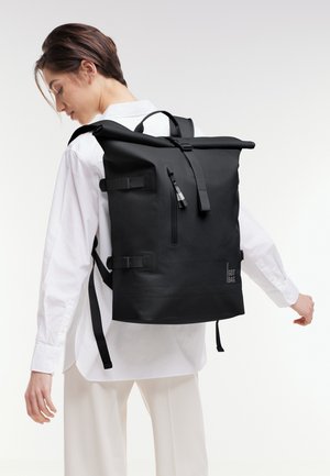 Junge Person mit großem schwarzem Rolltop-Rucksack mit Front-Reißverschlusstasche, gekleidet in weißem Hemd und heller Hose, steht vor weißem Hintergrund.