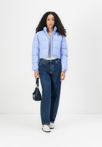 Lys lilla kort pufferjakke med kviltet mønster, hvit topp, høyt liv blå jeans, og en liten svart håndveske.