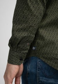 Camisa de manga larga verde con un diseño geométrico negro, con puños abotonados y una etiqueta en el dobladillo. Llevada con jeans azules.