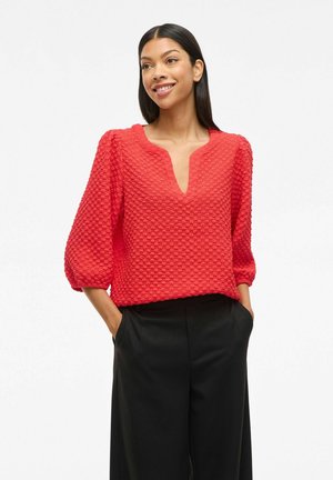 Woman wearing textured red V-neck blouse with puff sleeves and black wide-leg pants, hands in pockets, smiling slightly, against white background.