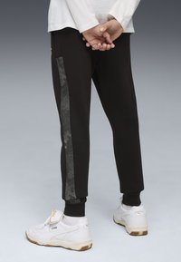 Pantalones de chándal negros con una franja lateral texturizada gris, puños elásticos y un corte relajado, combinados con zapatillas deportivas blancas.