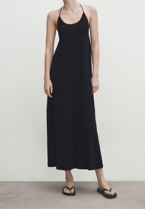 Femme portant une longue robe noire à fines bretelles et des sandales noires à semelles plates, debout devant un fond clair uni.