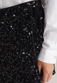 Juodas blizgučių delninukas su tekstūruota paviršiumi; turi švelnų, aksominį juostelės užsegimą ir glotnų audinio pamušalą. Laikomas rankoje, matoma ranka.