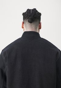 Czarna kurtka jeansowa z kontrastującym czarnym dzianinowym kołnierzem, widocznymi detalami szwów oraz teksturowanym wyglądem, widziana od tyłu.