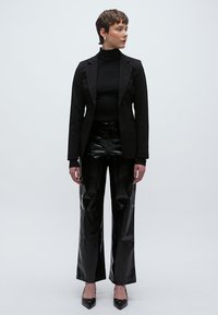 Fekete, testhezálló blézer fekete, testhez simuló garbó fölött, magasderekú, fényes fekete, bővülő szárú nadrággal és hegyes orrú fekete magassarkúval párosítva.