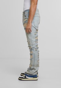 Šviesiai mėlyni nuvaryti džinsai su keliais dideliais skersiniais ir suplėšytais kraštais. Džinsai yra lieso kirpimo, derinami su juodais ir baltais sportbačiais.