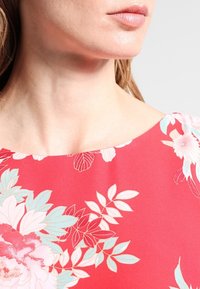 Blouse florale avec un fond rouge vif, de grands motifs de fleurs bleu clair et blancs, et une texture de tissu lisse et douce.