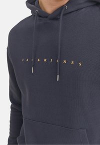 Marineblauer Hoodie aus strukturiertem Stoff, mit einer Vorderentasche, Kordeln und goldenen Schriftzug "JACK & JONES" auf der Brust.