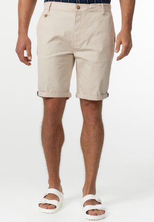 Homme portant un short beige retroussé, une chemise bleue à motifs rentrée dans le pantalon et des sandales blanches à double bride, debout devant un fond blanc.