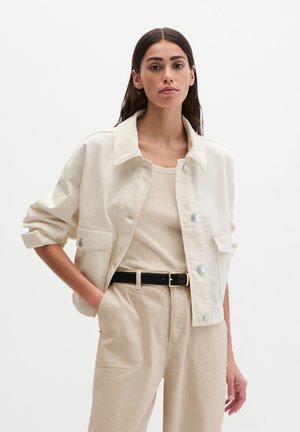 Femme portant une veste courte crème par-dessus un débardeur côtelé beige, un pantalon beige taille haute avec ceinture noire, debout devant un fond blanc.