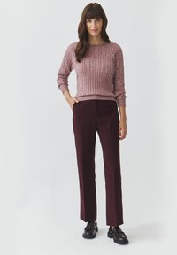 Cable-knit pink sweater with crew neck and long sleeves, paired with high-waisted burgundy trousers. Model wears black loafers.