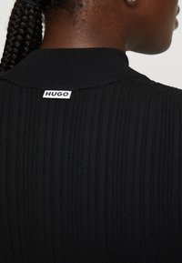 Black ribbed pullover featuring a high collar and a white "HUGO" tag at the back. The fabric appears textured and form-fitting.