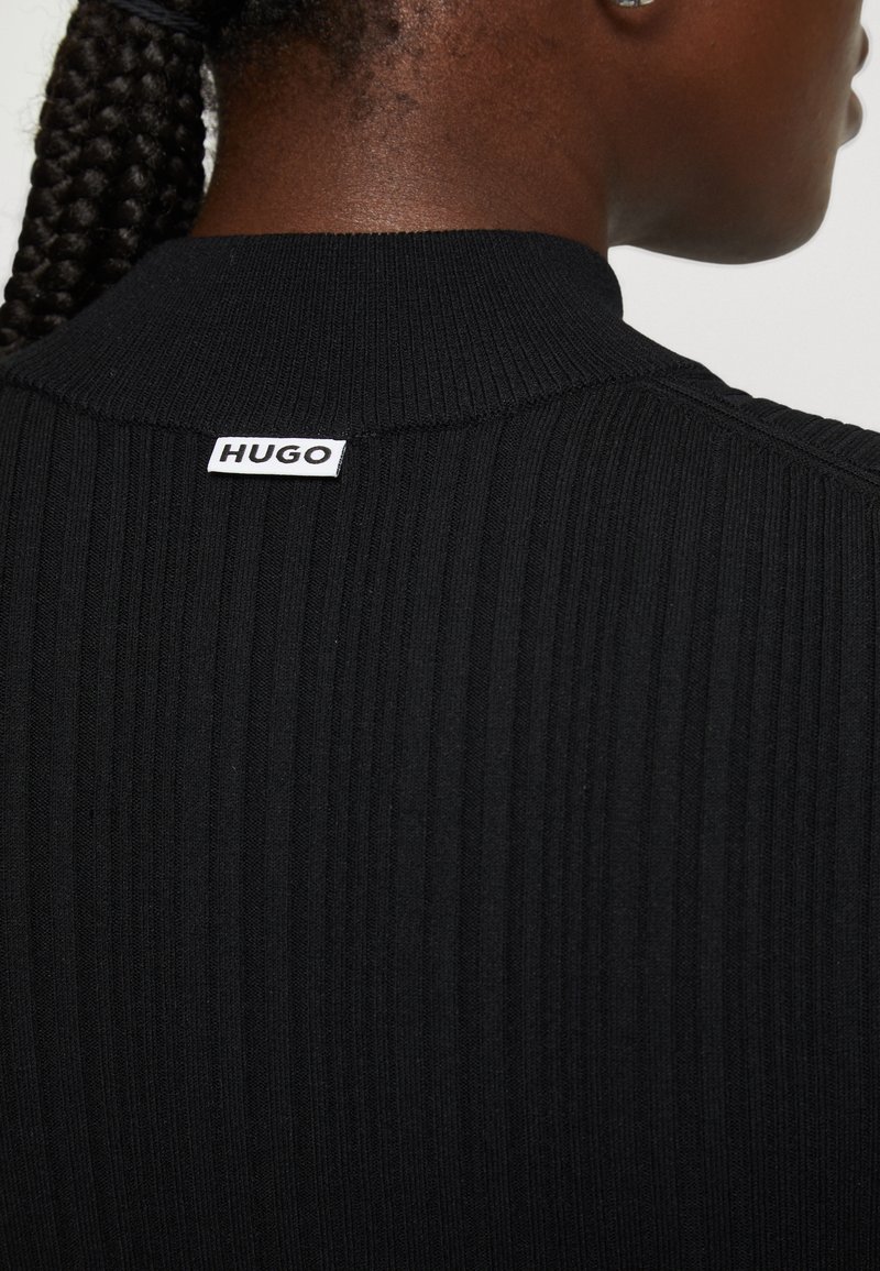 Schwarzer, gerippter Pullover mit hohem Kragen und einem weißen "HUGO"-Etikett auf der Rückseite. Der Stoff wirkt strukturiert und körpernah.