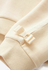 Poignet de sweat-beige avec texture côtelée, comportant un petit nœud en ruban crème et une étiquette logo blanche discrète le long de la couture.