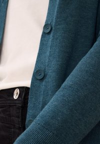 Teal cardigan met een gestructureerde brei, voorzien van vijf donkere knopen. Gedragen over een wit shirt en zwarte broek, wat een gelaagd uiterlijk benadrukt.