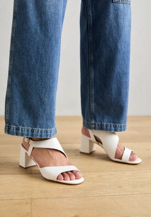 Witte leren sandalen met blokhak en open teen, met kruiselings banden en een enkelriempje met gesp. Gecombineerd met wijde blauwe spijkerbroek.
