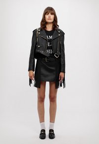 Giacca da moto in pelle nera con accessori dorati, abbinata a una gonna nera con frange e mocassini neri. Modella in piedi su uno sfondo bianco.