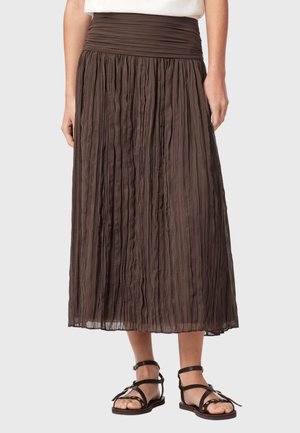 Brun plisseret midi-nederdel med bred talje, båret med sorte flade sandaler med ankelsnor på en person mod en ensfarvet baggrund.