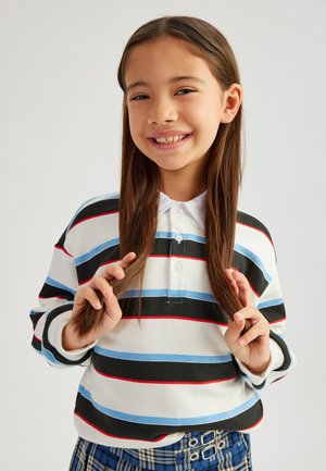 Gestreiftes Polo in Weiß und Schwarz mit blauen und roten Linien; geschichteter Kragen; das Model hat lange braune Haare und trägt einen karierten Rock mit Schnallendetails.