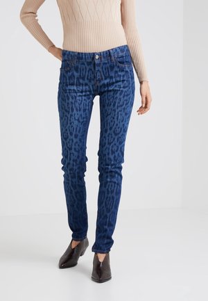 Mujer con vaqueros ajustados de estampado de leopardo azul, camiseta de manga larga beige con textura acanalada y botines negros con punta, posando sobre un fondo blanco.