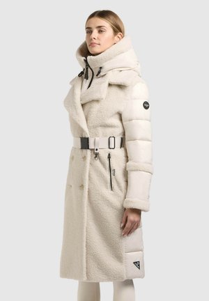 Femme portant un long manteau en peau de mouton et matelassé blanc cassé, avec un col montant et une taille ceinturée, debout sur un fond uni.