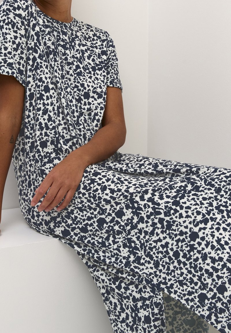 Personne portant une robe blanche avec des taches abstraites noires, assise avec un bras appuyé sur une surface blanche et l’autre sur ses genoux.