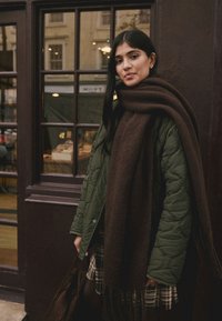 Quilted green coat, brown wool scarf draped, checkered dress, dark tights, brown handbag, standing by windows with a wooden frame.