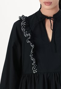 Μαύρο φόρεμα με μακριά μανίκια, που διαθέτει βαθύ V-λαιμόκοψη, ριχτό κρόσσι στους ώμους και περίπλοκη λευκή κεντητική γύρω από τον κρόσσο.