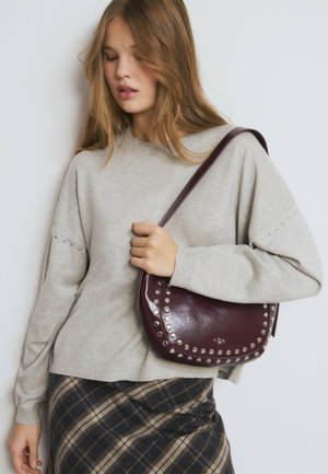 Sac à main rond en cuir bordeaux avec des clous argentés le long des bords. L'accessoire contraste avec un pull oversize gris clair et une jupe à carreaux.