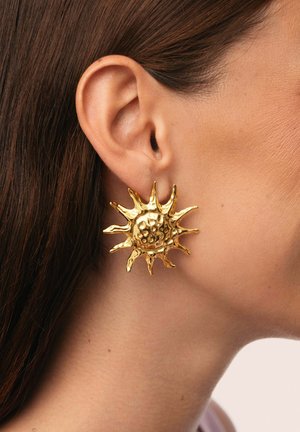 Pendientes en forma de sol dorados con superficie texturizada y acentos puntiagudos, que exhiben un diseño tridimensional en el lóbulo de la oreja.